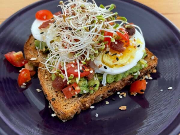 slice of toast topped with mashed avocado, sliced hard boiled egg, chopped cheery tomatoes, sprouts and crumbed bacon on a blue pottery plate