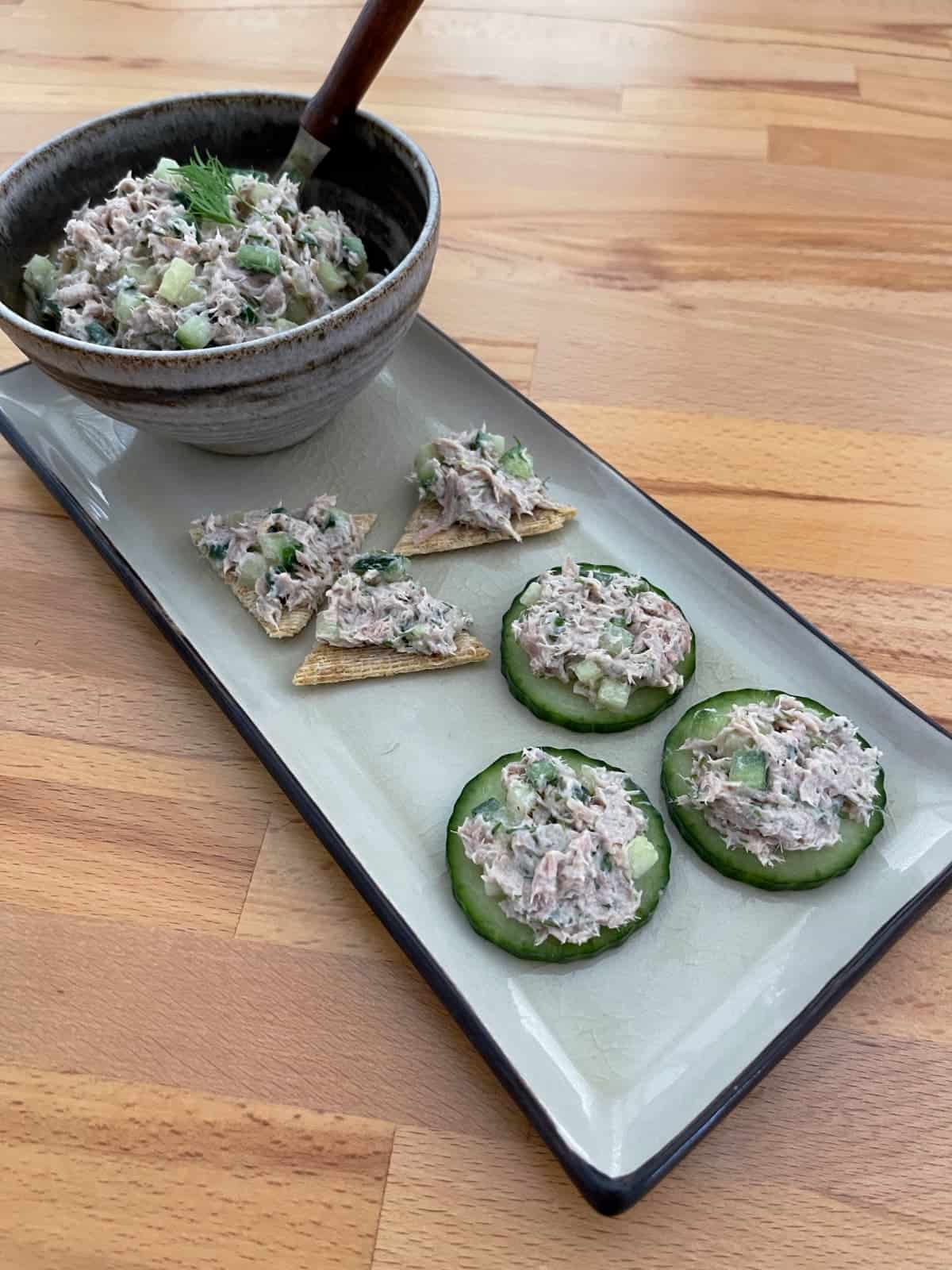 Dilly tuna spread in bowl on platter with cucumber slices and crackers topped with tuna.