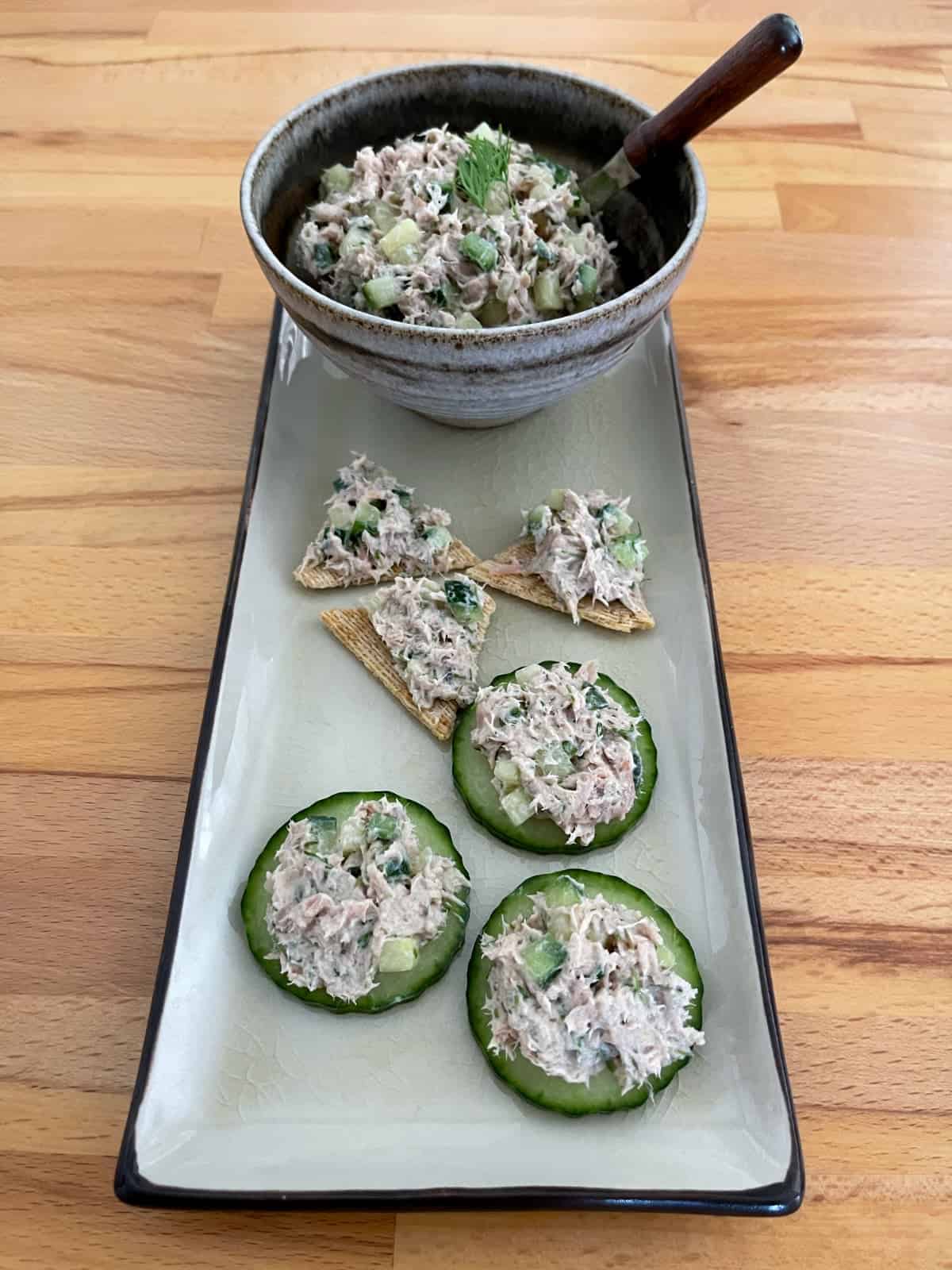 Tuna spread with fresh dill in small serving bowl with tuna topped crackers and cucumber slices on green platter.