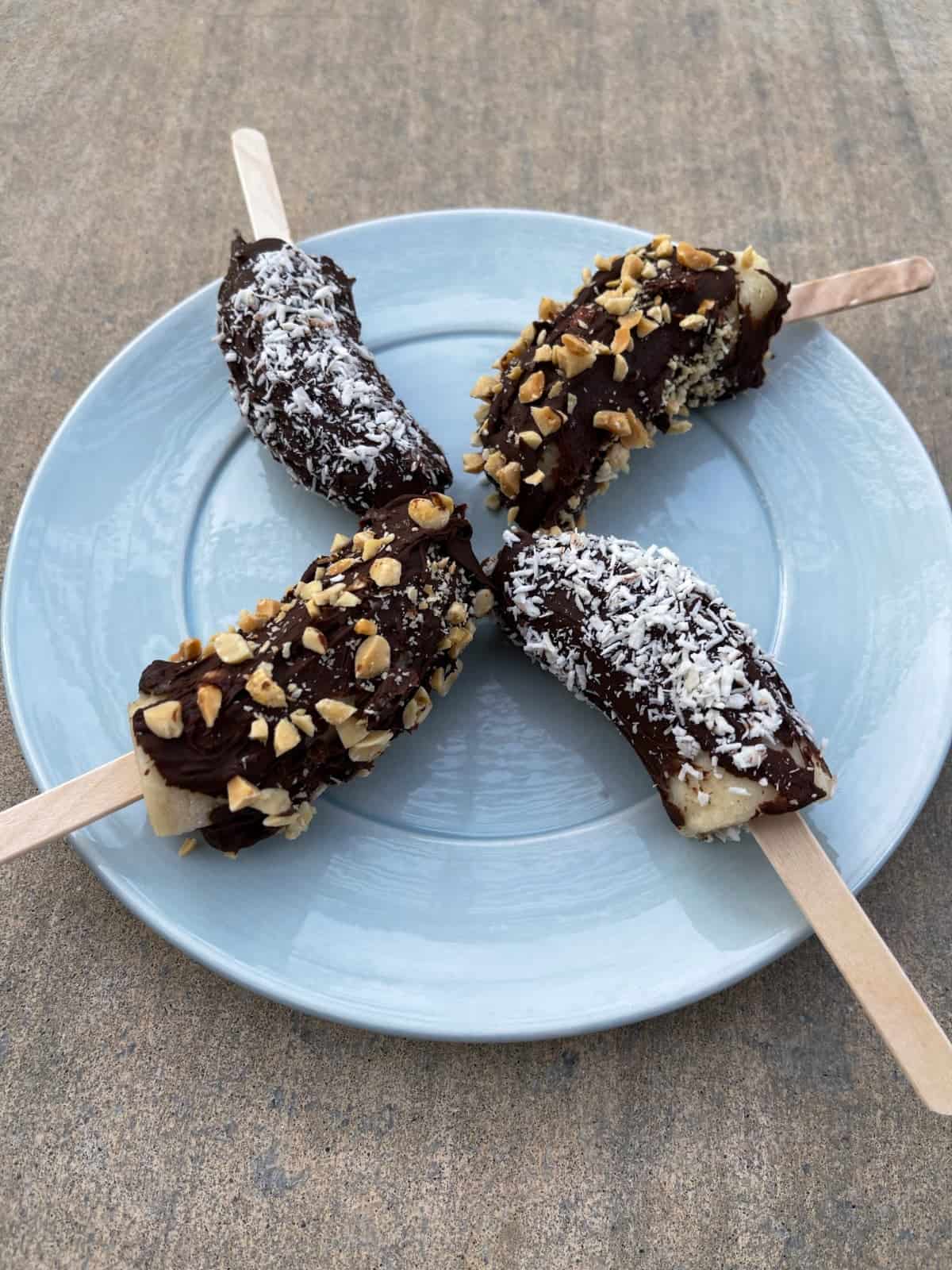 Four frozen chocolate bananas, two topped with shredded coconut and two with chopped peanuts, on a blue plate.