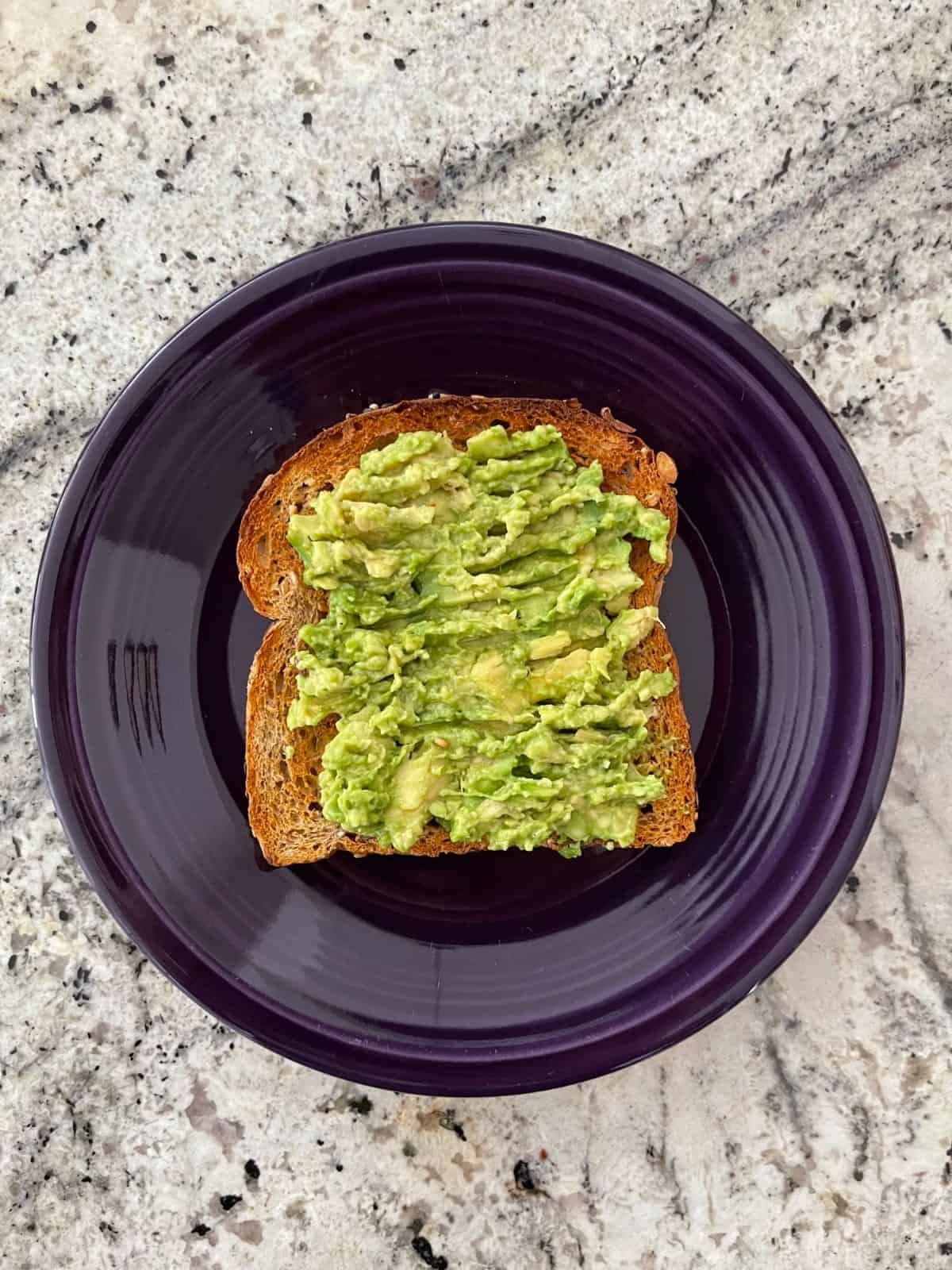 Mashed avocado on toasted whole grain bread on purple plate.
