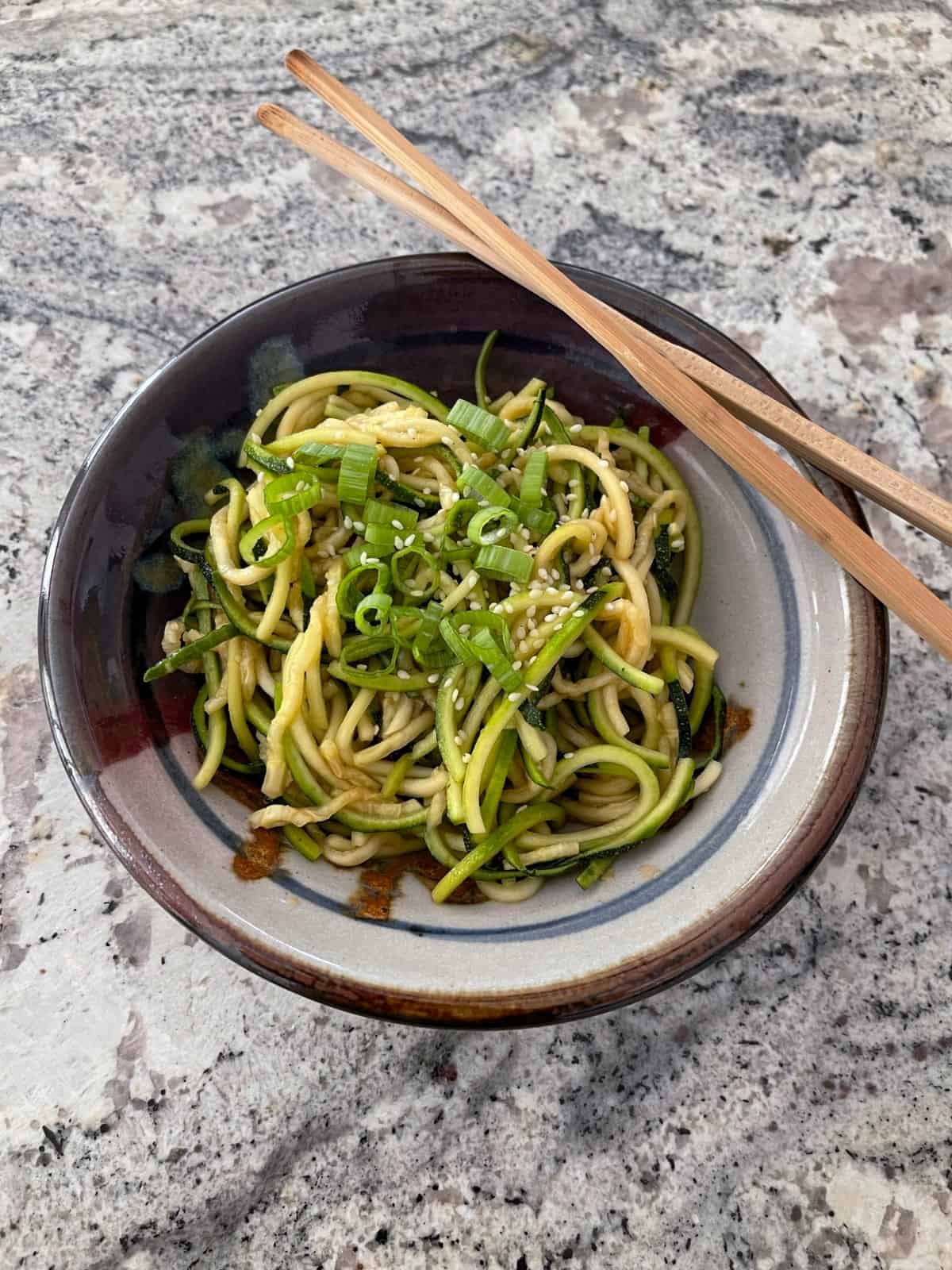 Sesame zucchini noodles garnished with green onion slices and sesame seeds in bowl with chop sticks.