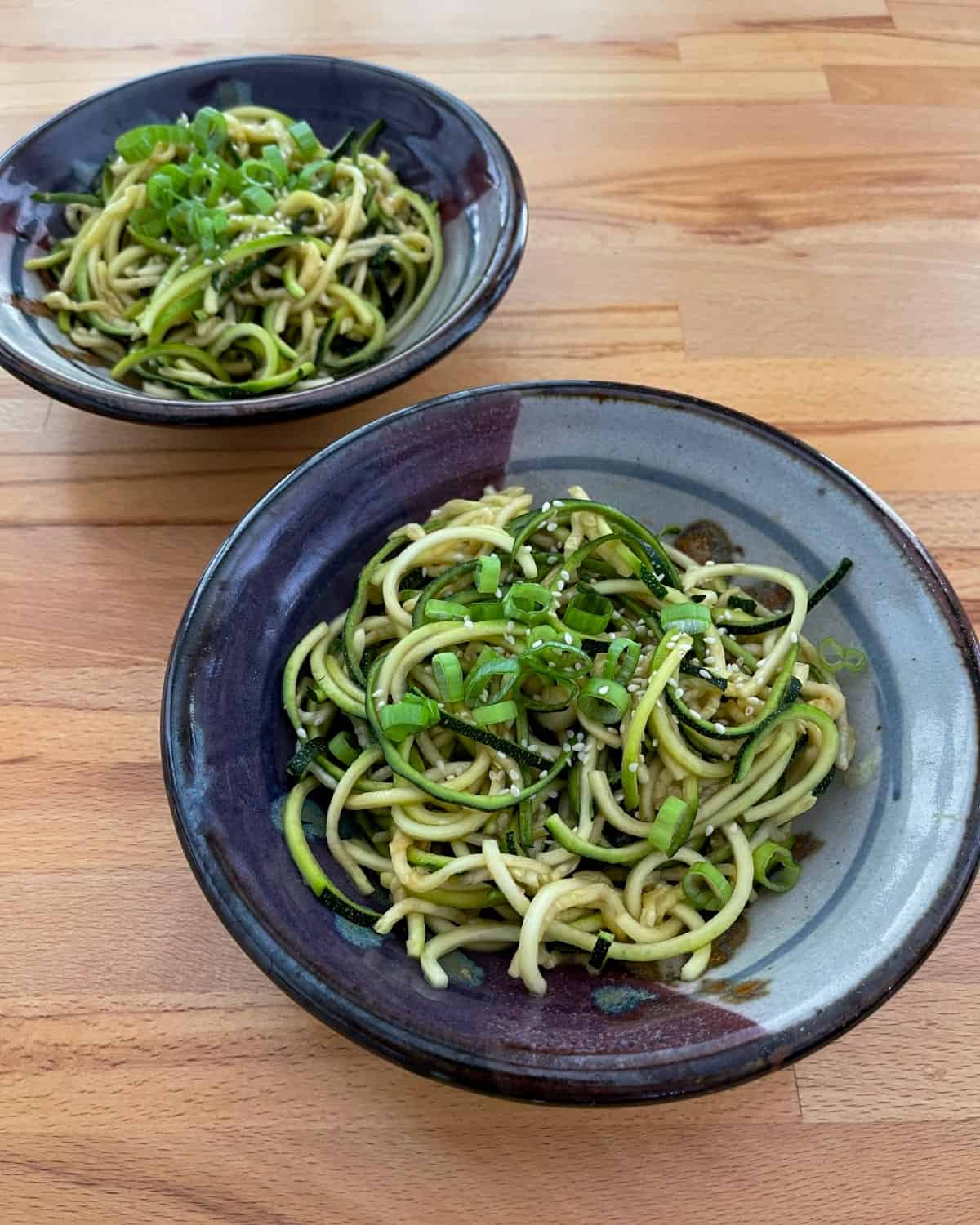 Two ceramic bowls with sesame zucchini noodles on wood table.