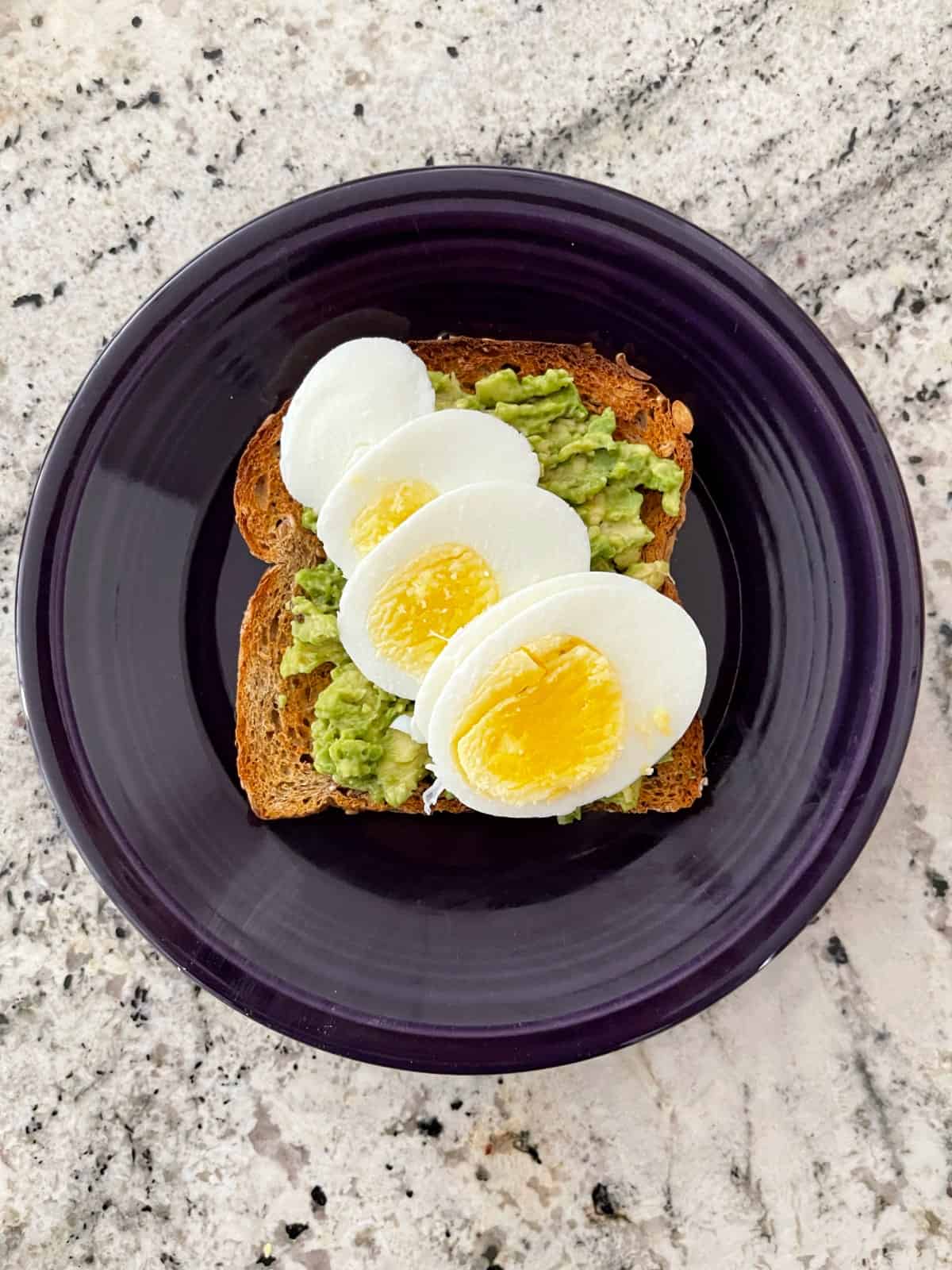 Avocado toast topped with sliced hard boiled egg on purple plate.