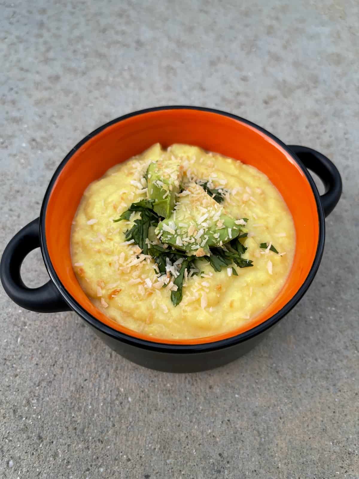 Chilled Coconut Corn Soup garnished with cilantro, avocado and toasted coconut in orange soup crock.