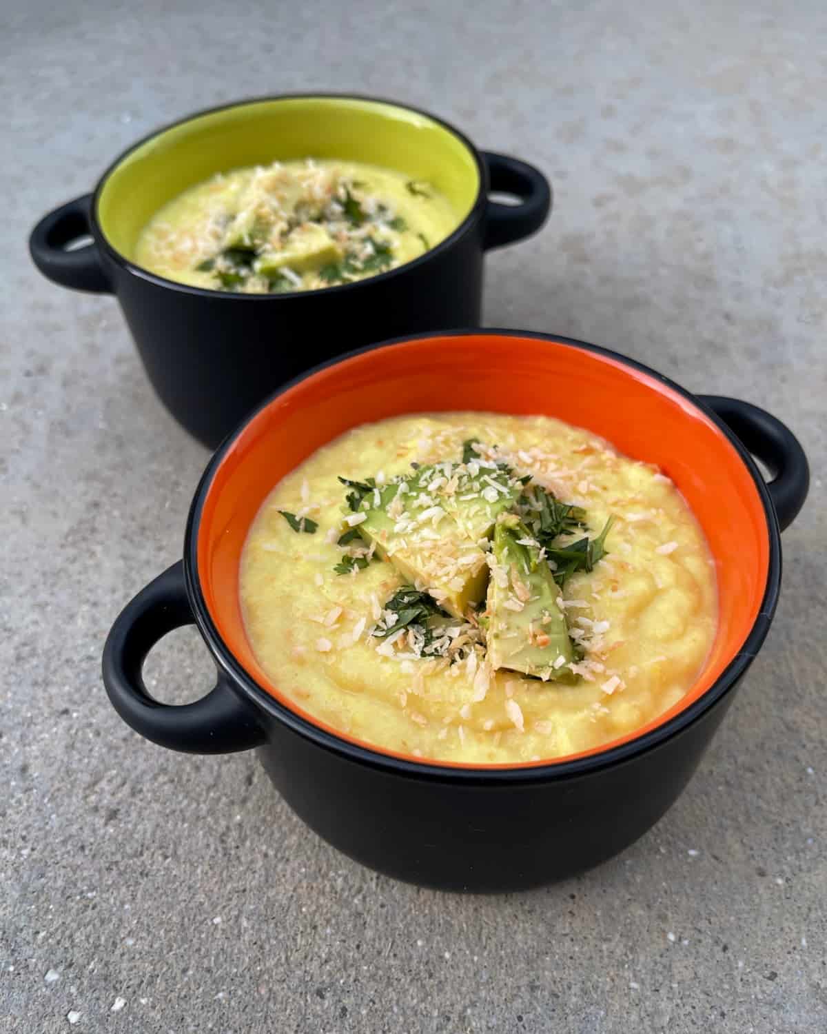 Two small bowls of chilled coconut corn soup topped with chopped avocado, cilantro and toasted coconut.