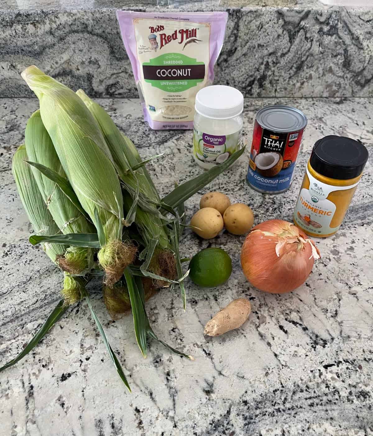 Ingredients for making coconut corn soup including corn on the cob, shredded coconut, coconut oil, coconut milk, potatoes, onion, ginger and lime.
