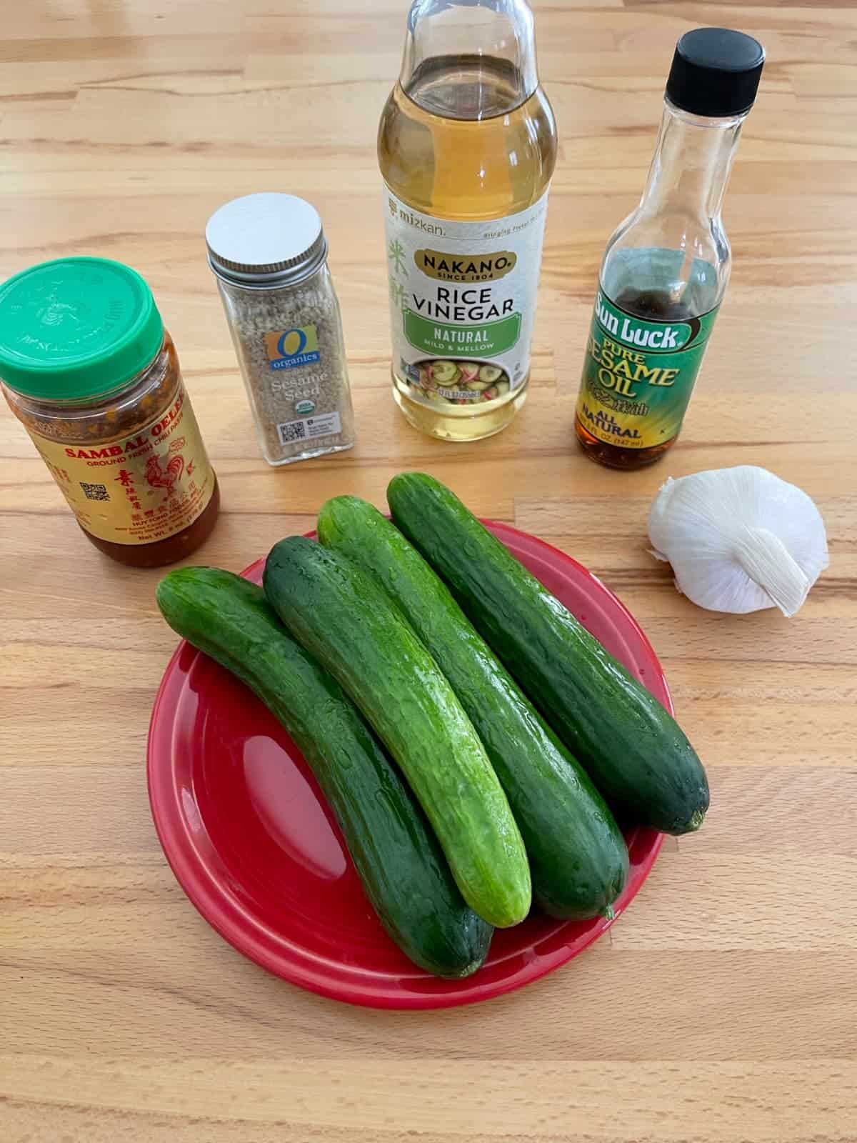 Ingredients for making spicy cucumber salad including mini cucumbers, sesame seeds, sesame oil, rice vinegar, garlic and Sambal oelek.