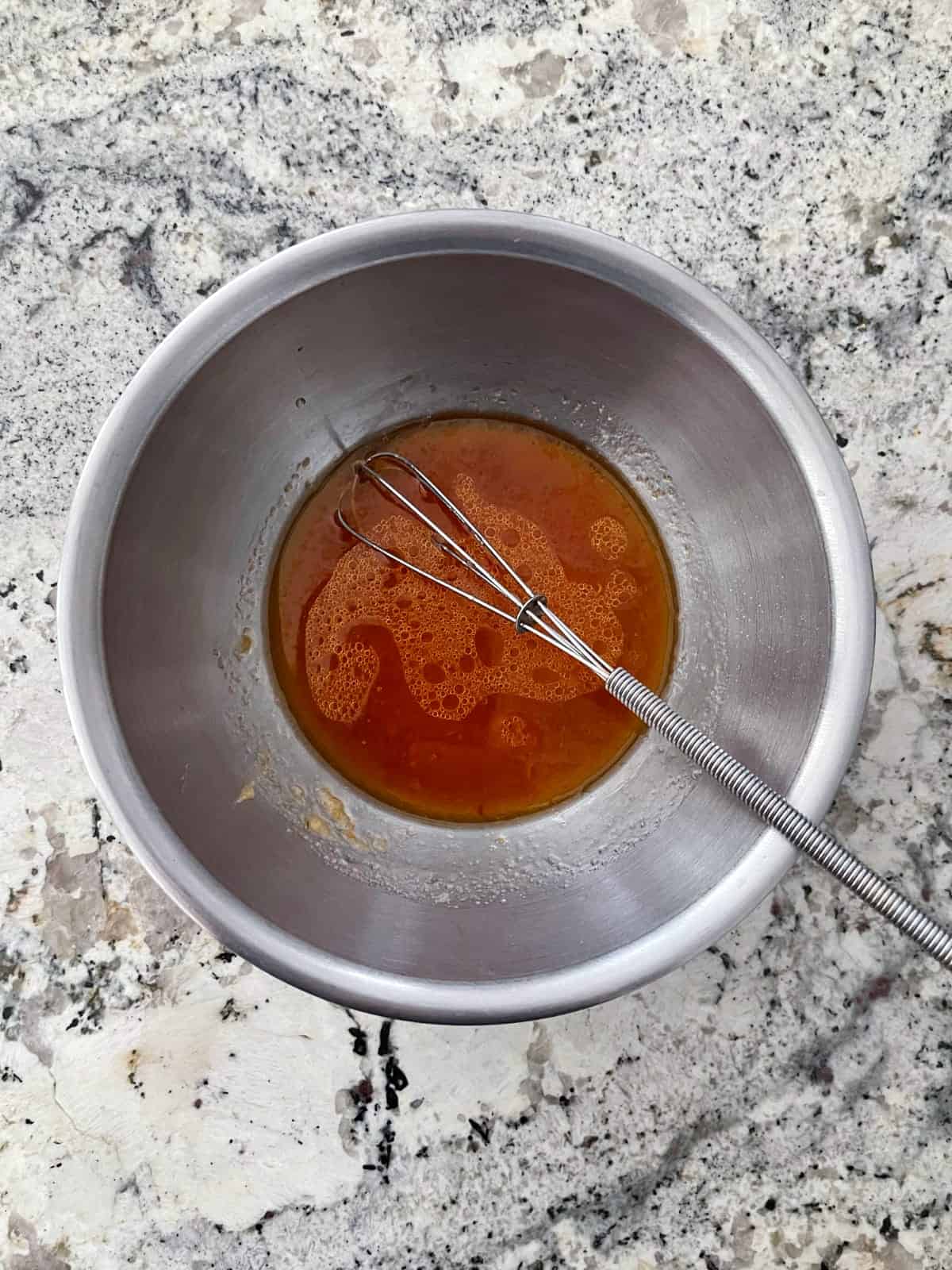 Whisking rice vinegar, Sambal oelek, sesame oil and garlic in stainless mixing bowl on granite counter.