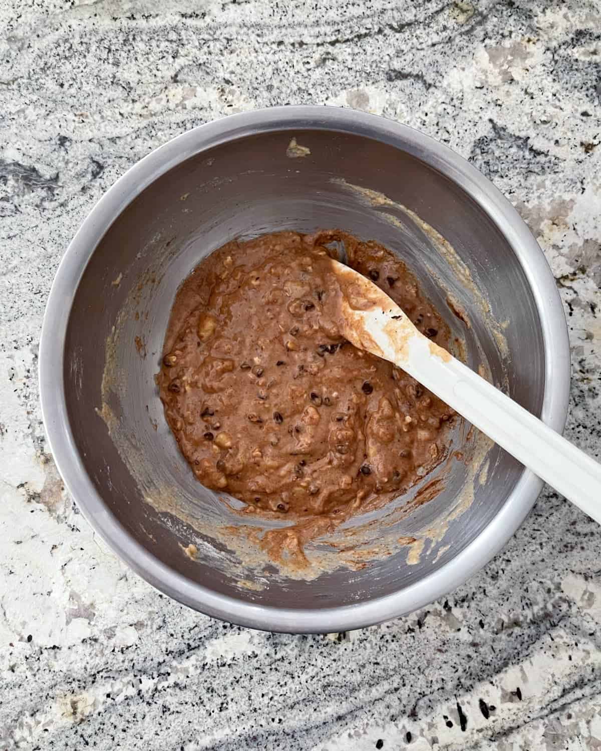Folding cocoa powder and mini chocolate chips into banana bread batter with spatula in mixing bowl.