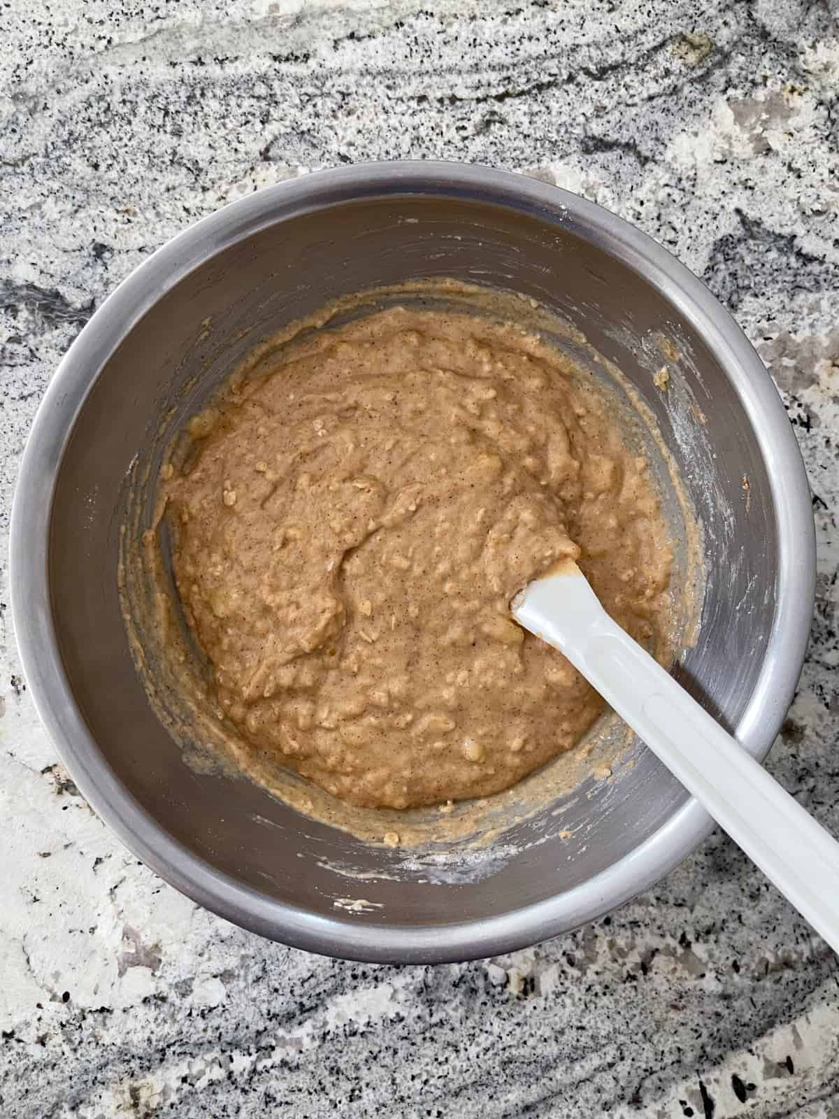 Folding oats and flour into mashed bananas, almond butter and oil in mixing bowl with spatula.