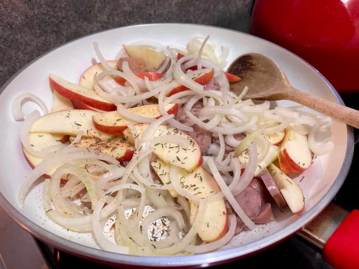 Apple slices, sliced onion and sausage in skillet with wooden spoon.
