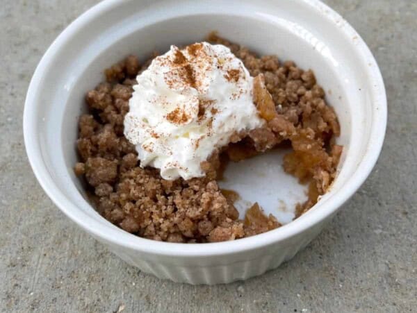 microwave apple crisp topped with whipped topping and sprinkle of cinnamon in white ceramic ramekin