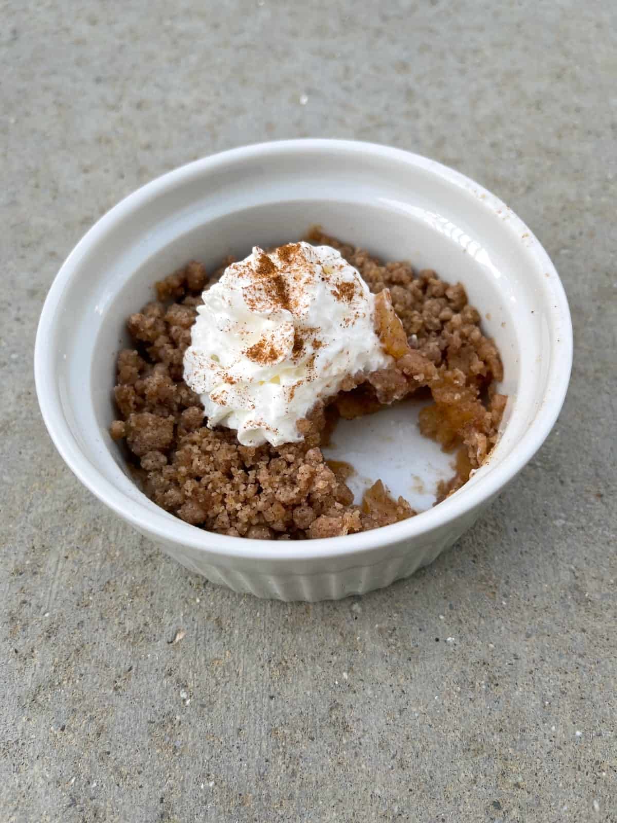 Partial microwave apple crisp dessert in white ramekin with whipped topping.