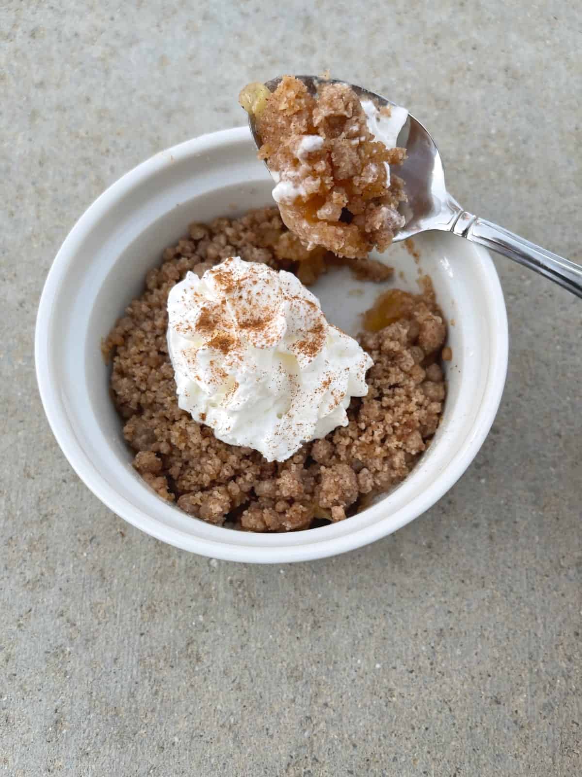 Microwave apple crisp in white ramekin with whipped topping and cinnamon sprinkle.