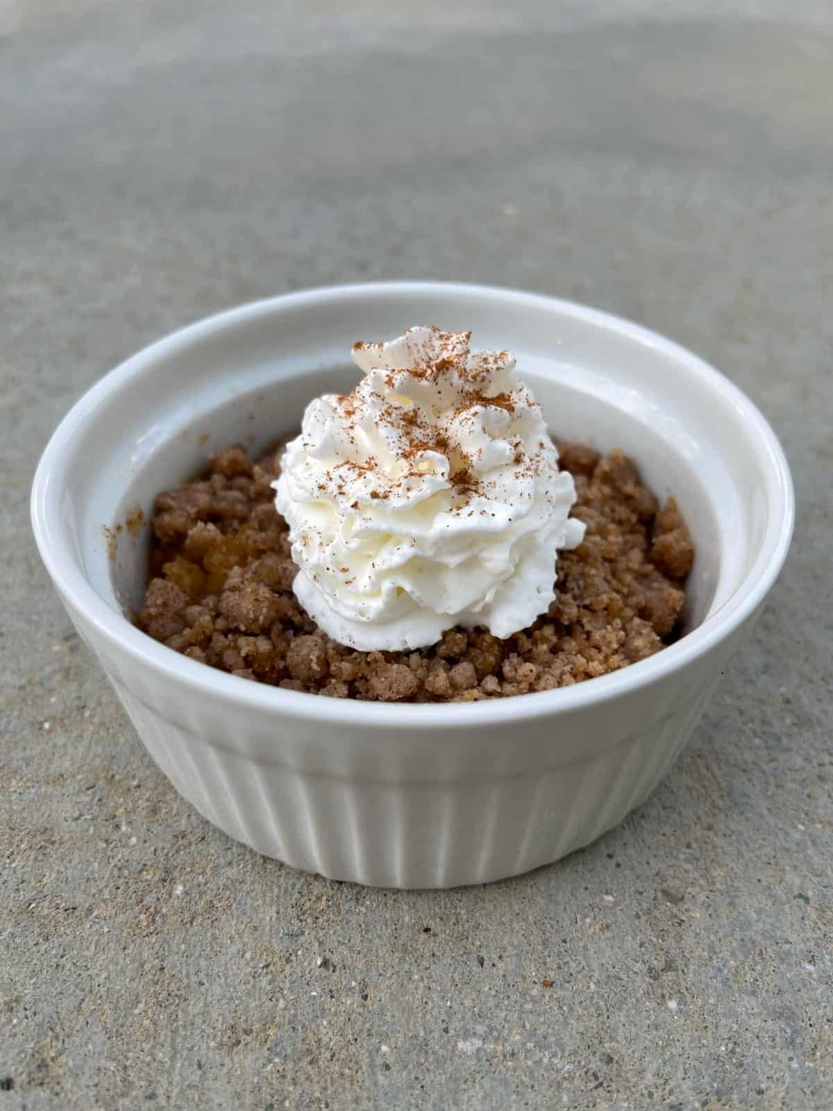 Microwave apple crisp in small white ramekin with whipped topping and cinnamon sprinkle.