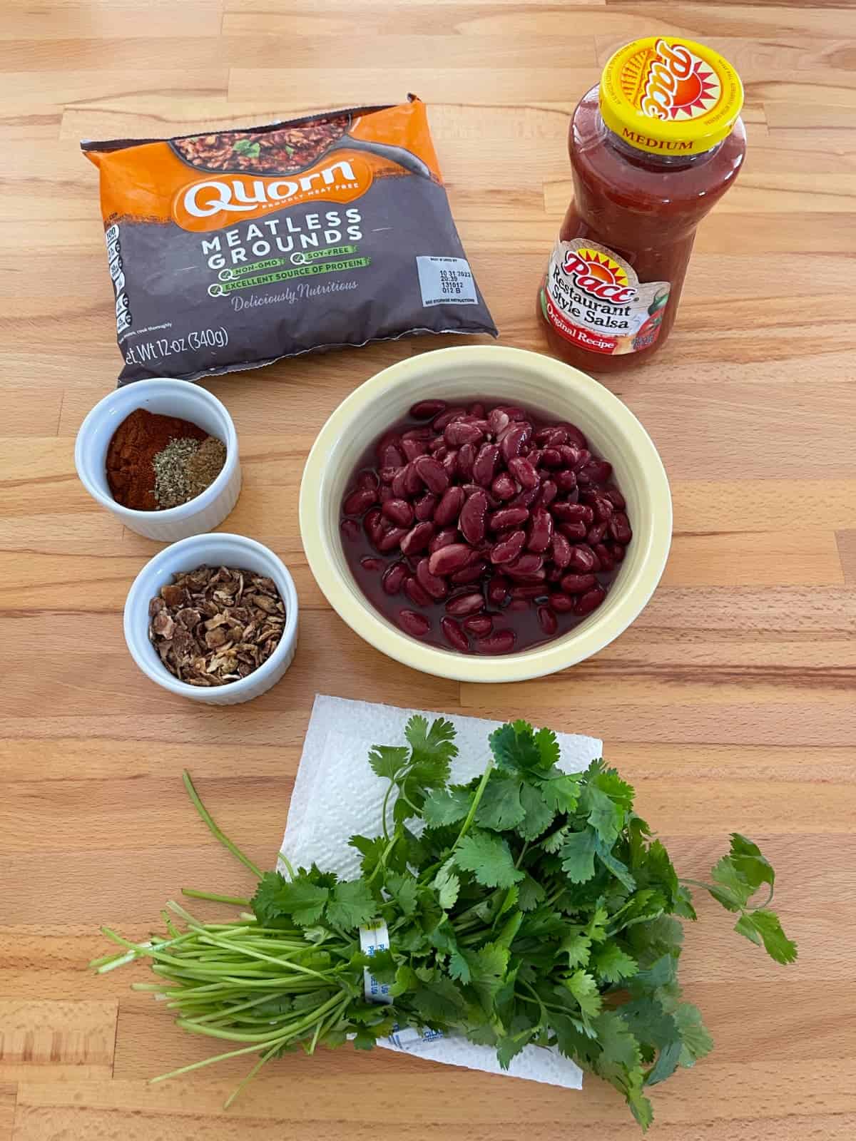 Ingredients for making chili, including Quorn meatless grounds, salsa, beans, spices and fresh cilantro on wood table.
