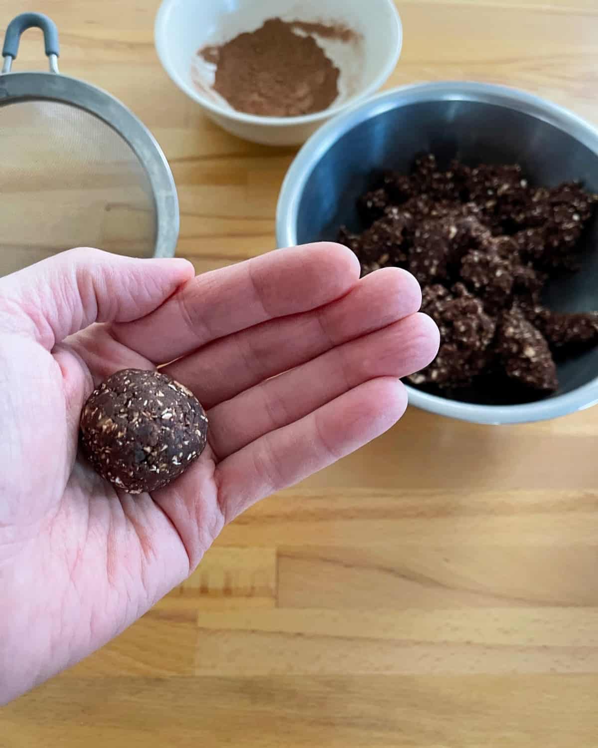 Rolling rum balls in hands with batter, cocoa powder and sieve in background.