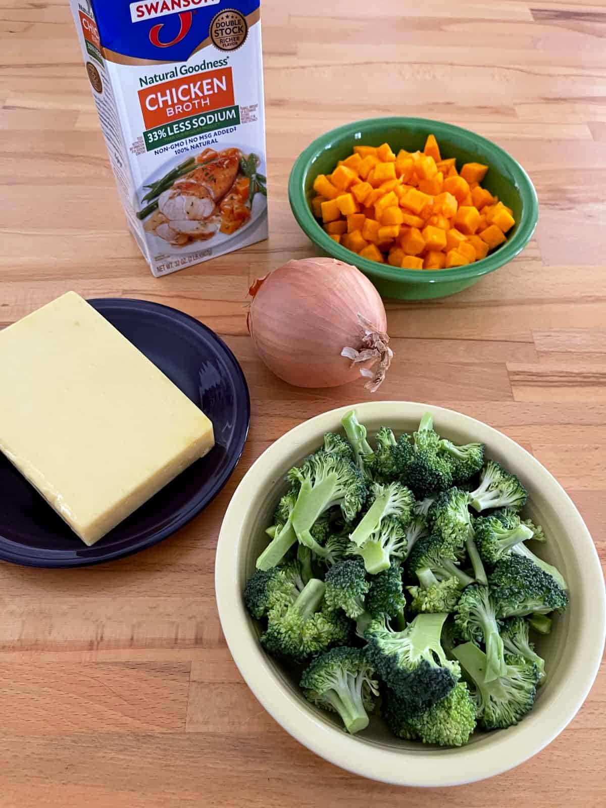 Ingredients for making soup, including broccoli florets, cheddar cheese, onion, butternut squash and chicken broth on wooden table.