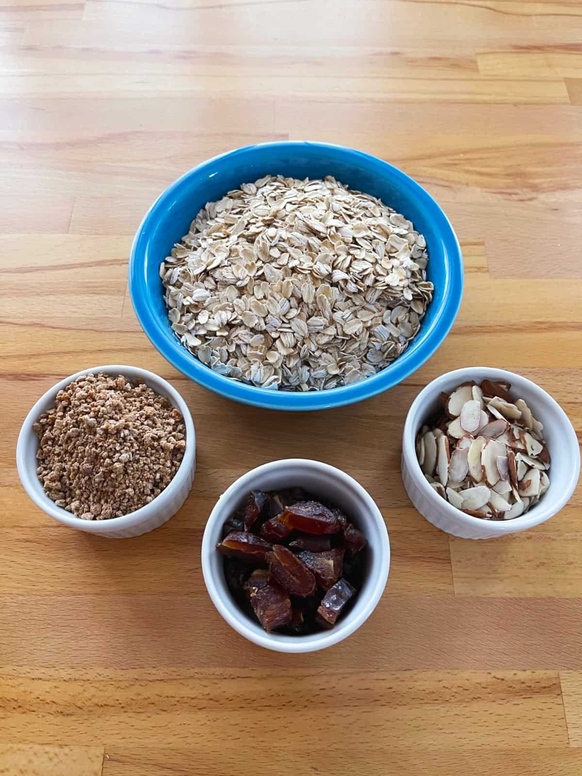 Ingredients for making oatmeal, including dry rolled oats, Grape Nuts, chopped date and sliced almonds on wooden table.