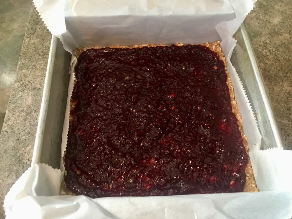 Spreading raspberry jam over oat crust in parchment lined baking pan.