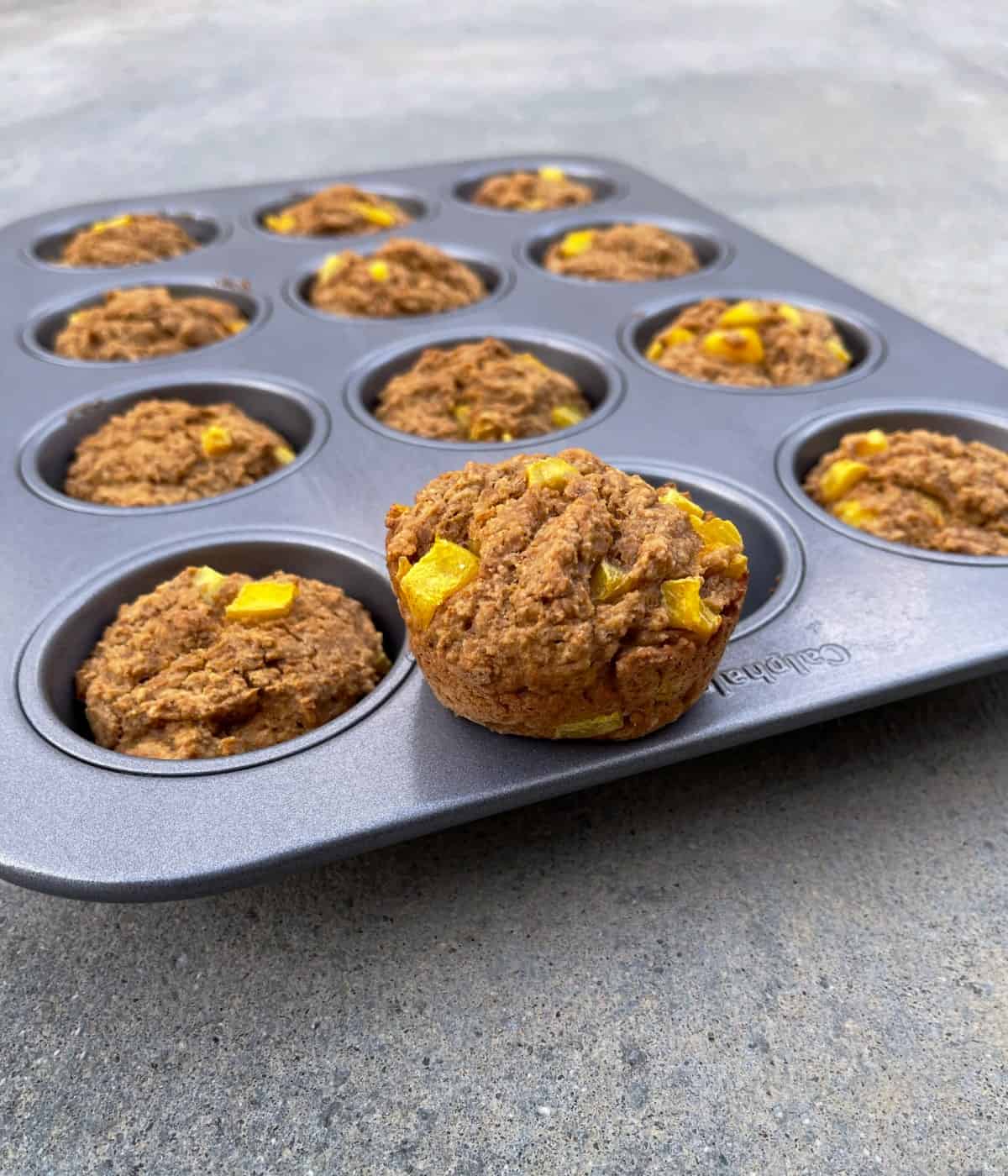 Fresh baked Fiber One mango muffins in muffin tin with one muffin on top of pan.