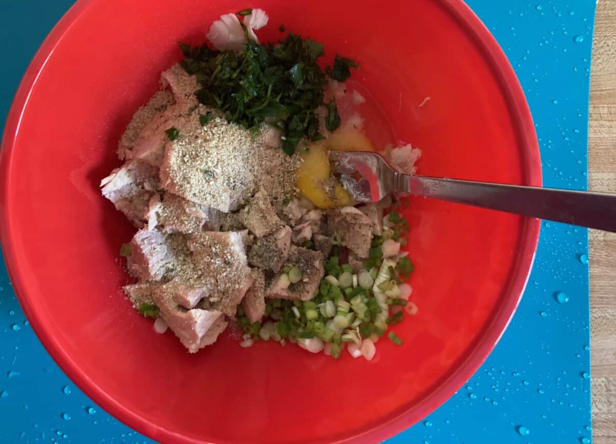 Mixing tuna, egg, bread crumbs, green onion and parsley in red bowl with a fork.