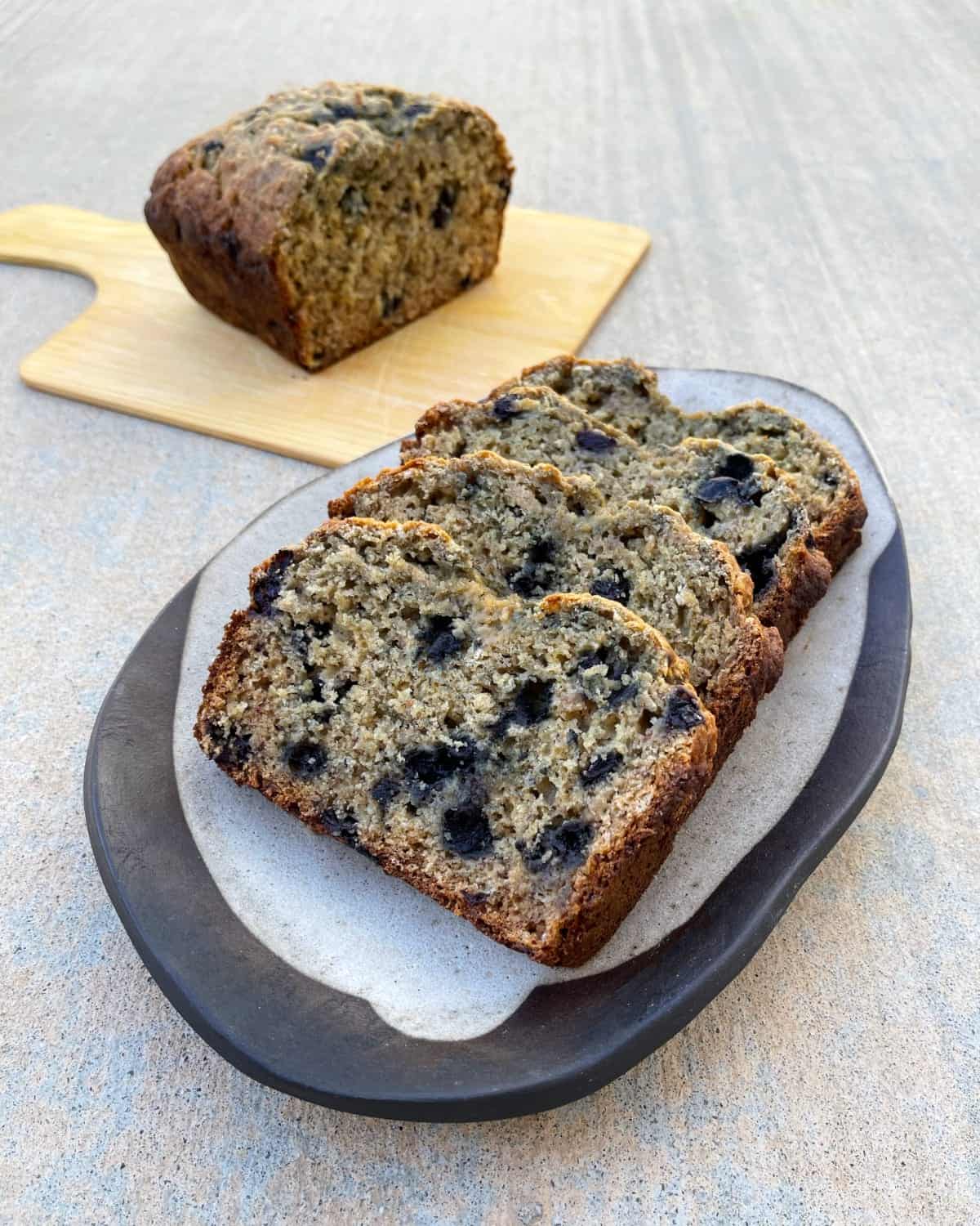 Blueberry banana bread slices on plate with half loaf on cutting board in background.