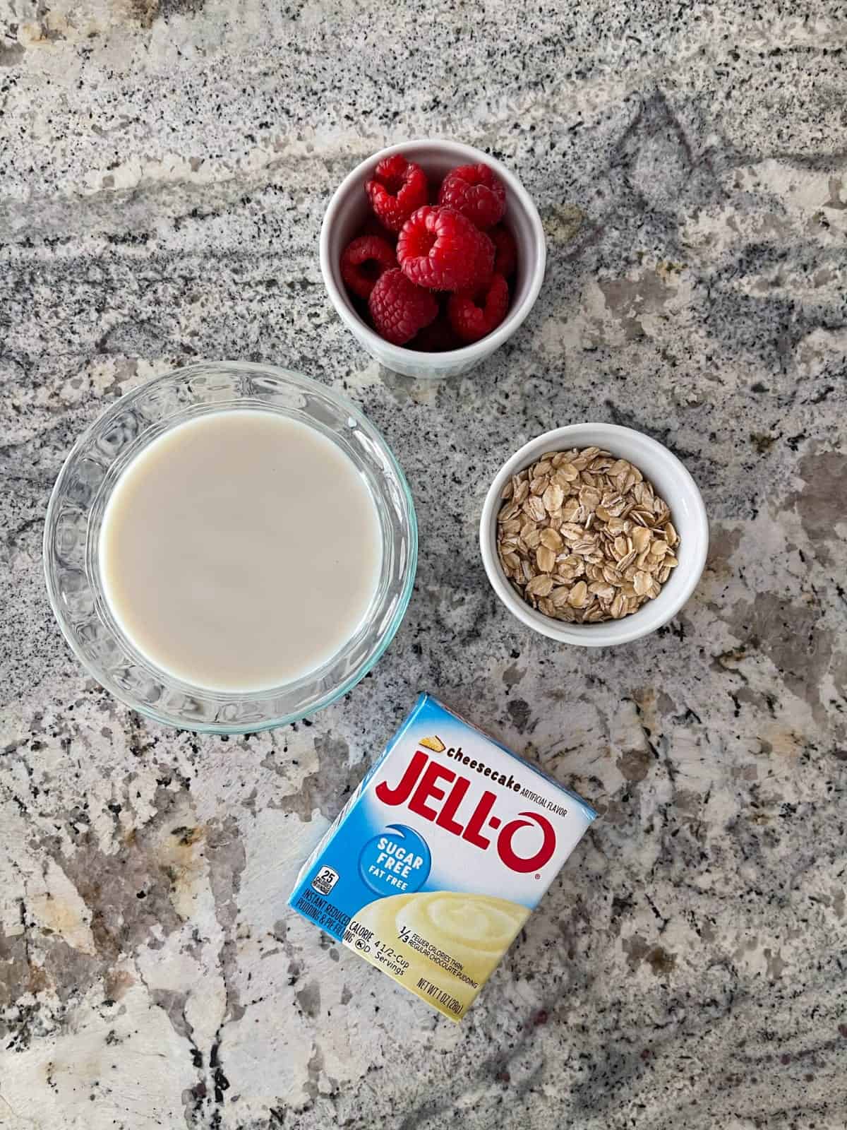 Ingredients on granite counter including sugar-free cheesecake Jello instant pudding mix, raspberries, oats and almond milk.