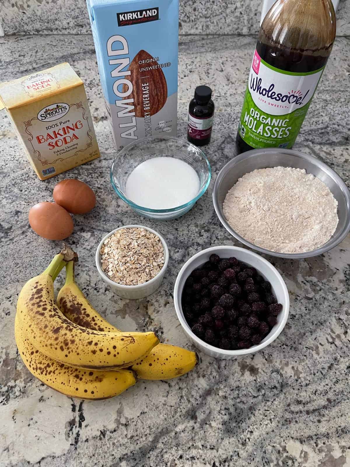 Ingredients for making blueberry banana bread including flour, blueberries, bananas, oats, eggs, molasses, sweetener and almond milk.