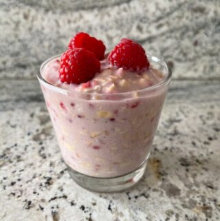 glass of raspberry cheesecake overnight oats topped with raspberries on granite countertop front view