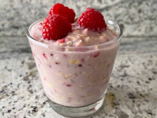 glass of raspberry cheesecake overnight oats topped with raspberries on granite countertop front view