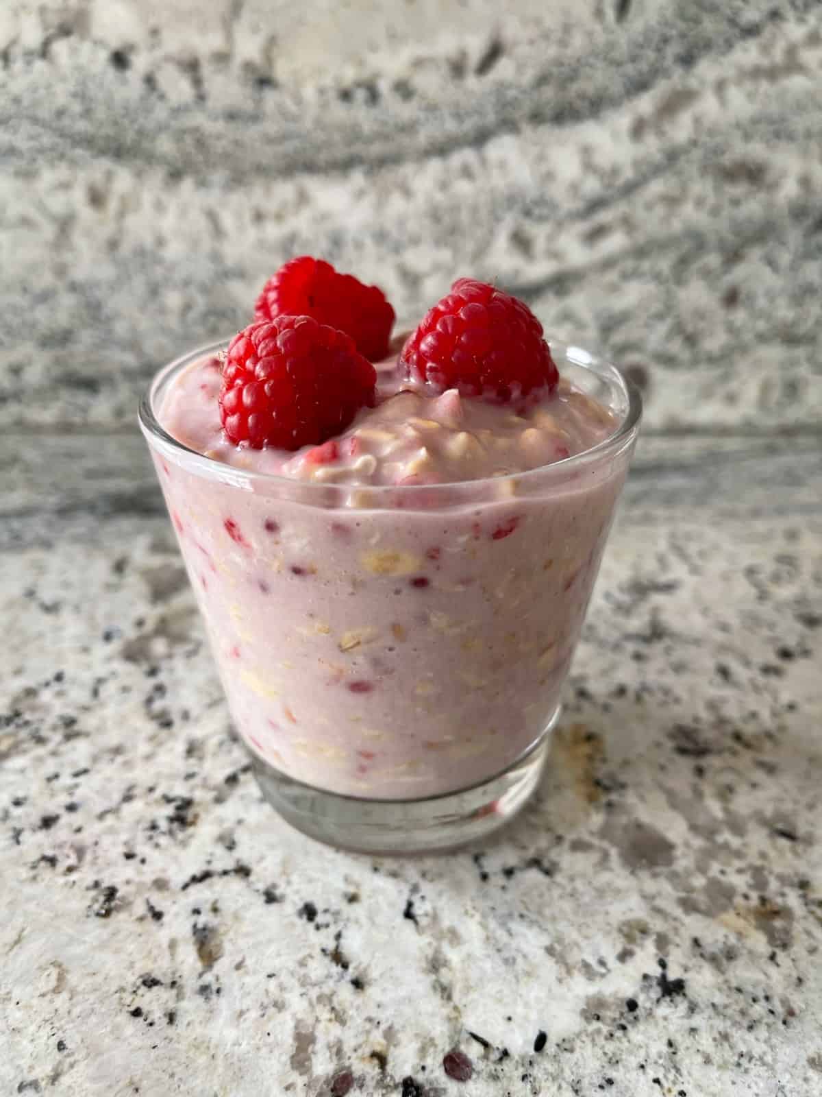 Glass of raspberry cheesecake overnight oats topped with raspberries on granite countertop.