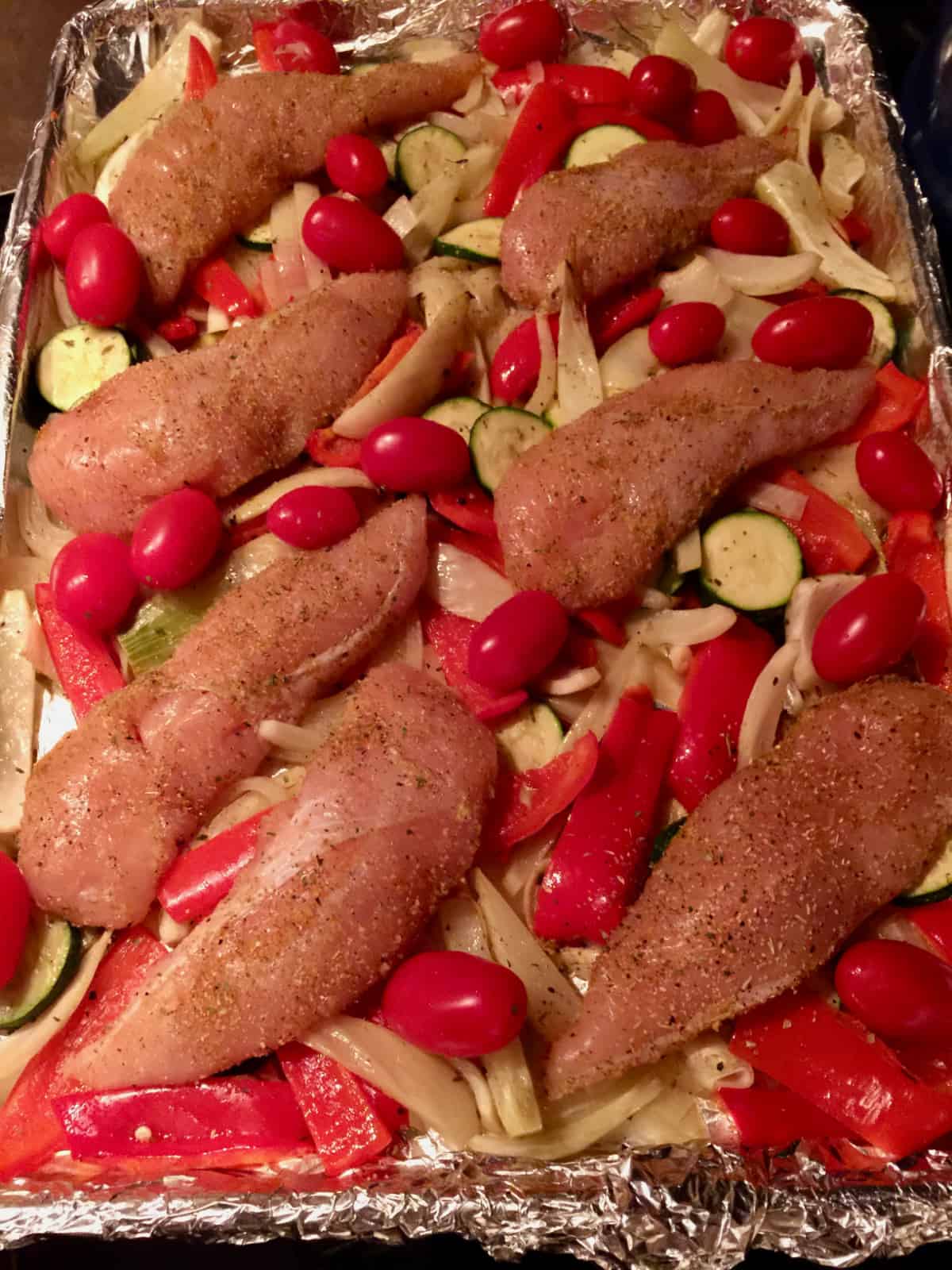 Foil-lined sheet pan with zucchini, onions, peppers, cherry tomatoes and chicken tenders.
