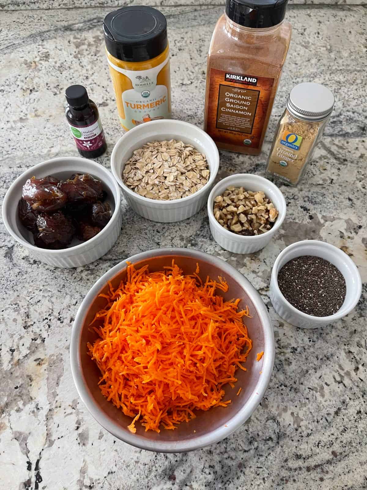 Ingredients including shredded carrots, dates, rolled oats, chopped walnuts, chia seeds, vanilla and spices for making carrot cake energy bites.