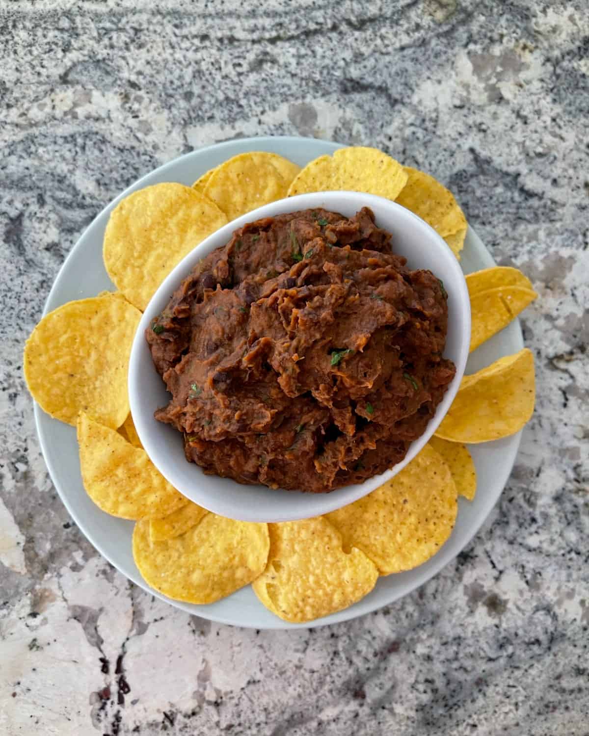 Refried black bean dip in white bowl with plate of yellow corn tortilla chips.