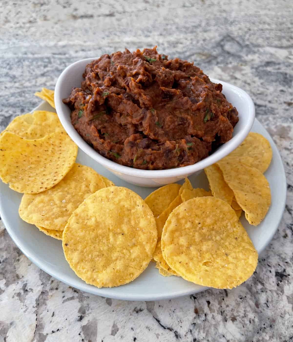 Warm refried black bean dip in white bowl with yellow corn tortilla chips.