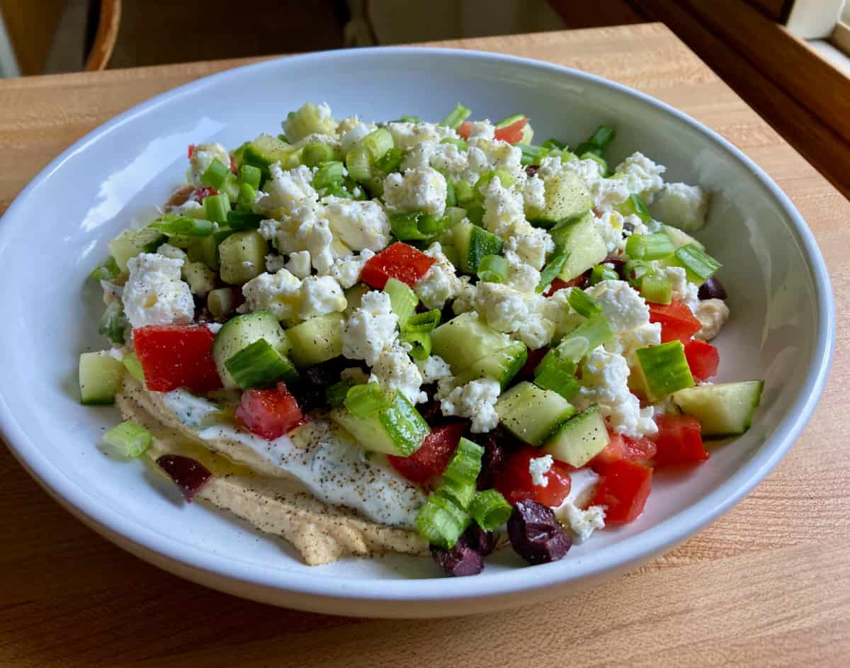 7 Layer Greek Dip with hummus, tzatziki, cucumbers and feta cheese in white serving bowl.