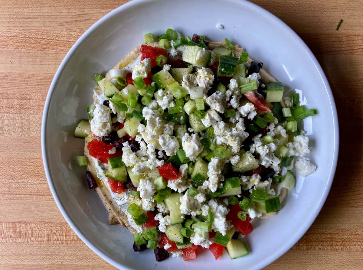 Layered Greek dip with hummus, tzatziki, cucumber, tomato, olives and feta cheese in white dish.