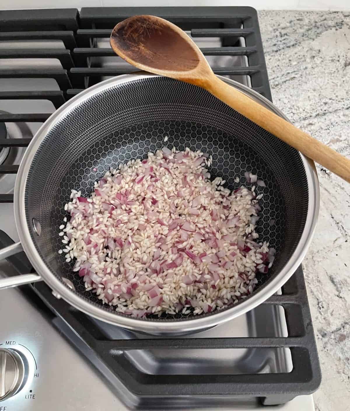 Arborio rice and chopped red onion in saucepan with wooden spoon.