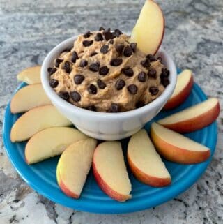 creamy peanut butter dip topped with chocolate chips in white cup surrounded by apple slices on a blue plate