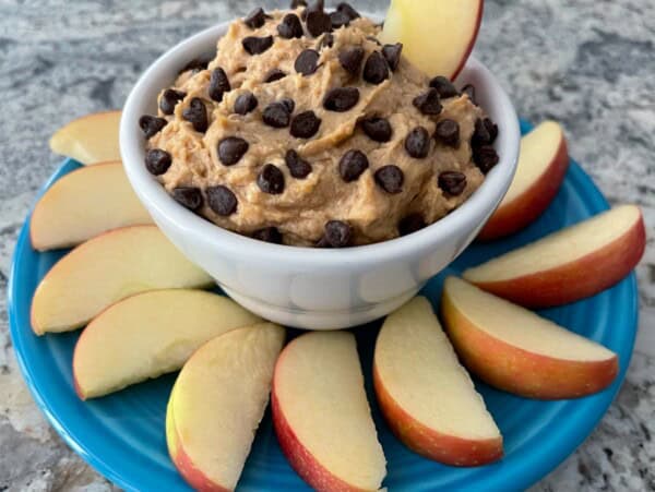 creamy peanut butter dip topped with chocolate chips in white cup surrounded by apple slices on a blue plate
