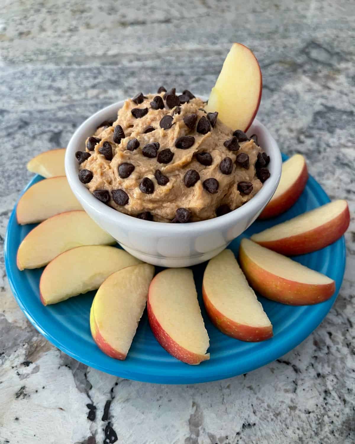 creamy peanut butter dip topped with chocolate chips in white cup surrounded by apple slices on a blue plate 