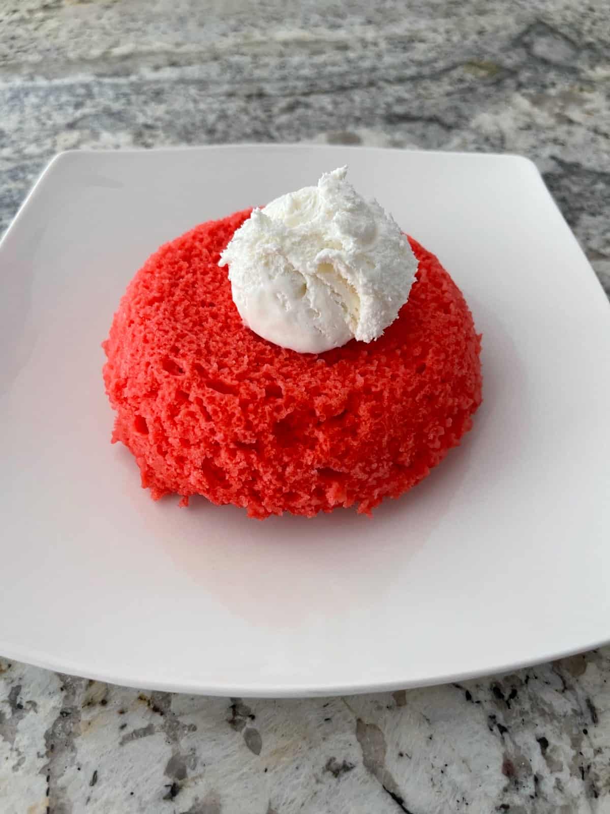 Raspberry Jell-o microwave cake topped with light whipped topping on small white plate.
