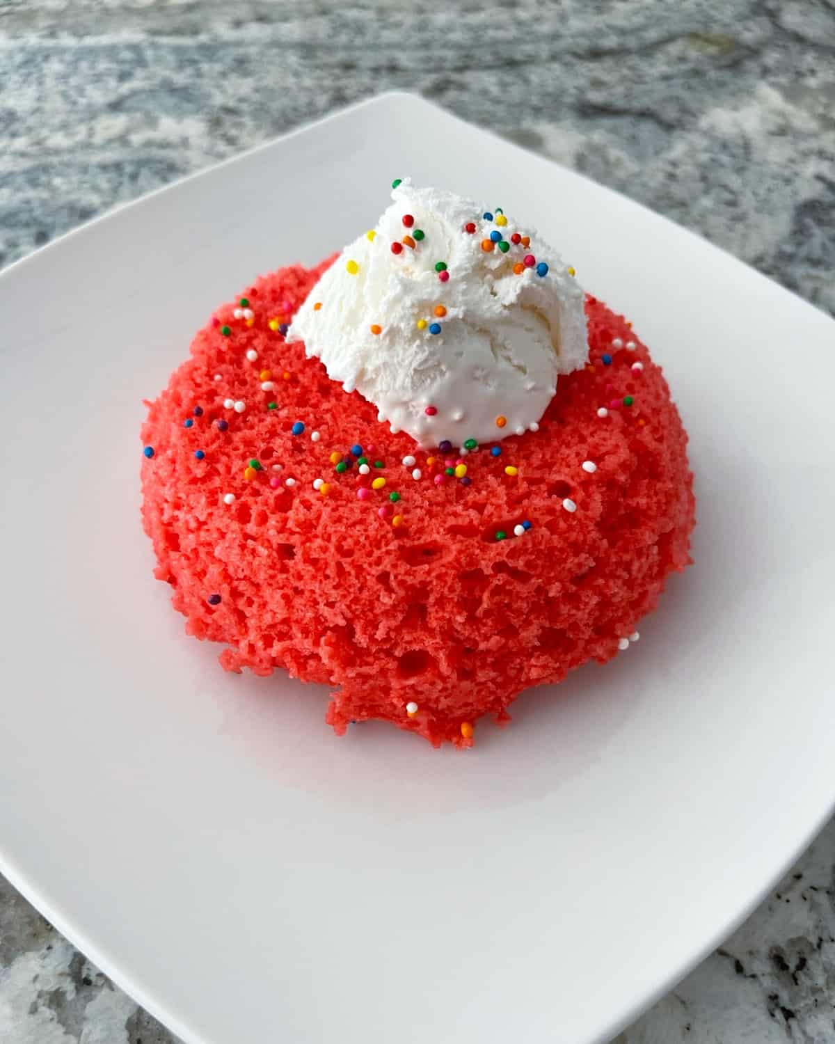 Raspberry Jell-o microwave mug cake topped with whipped topping and rainbow sprinkles on white plate.