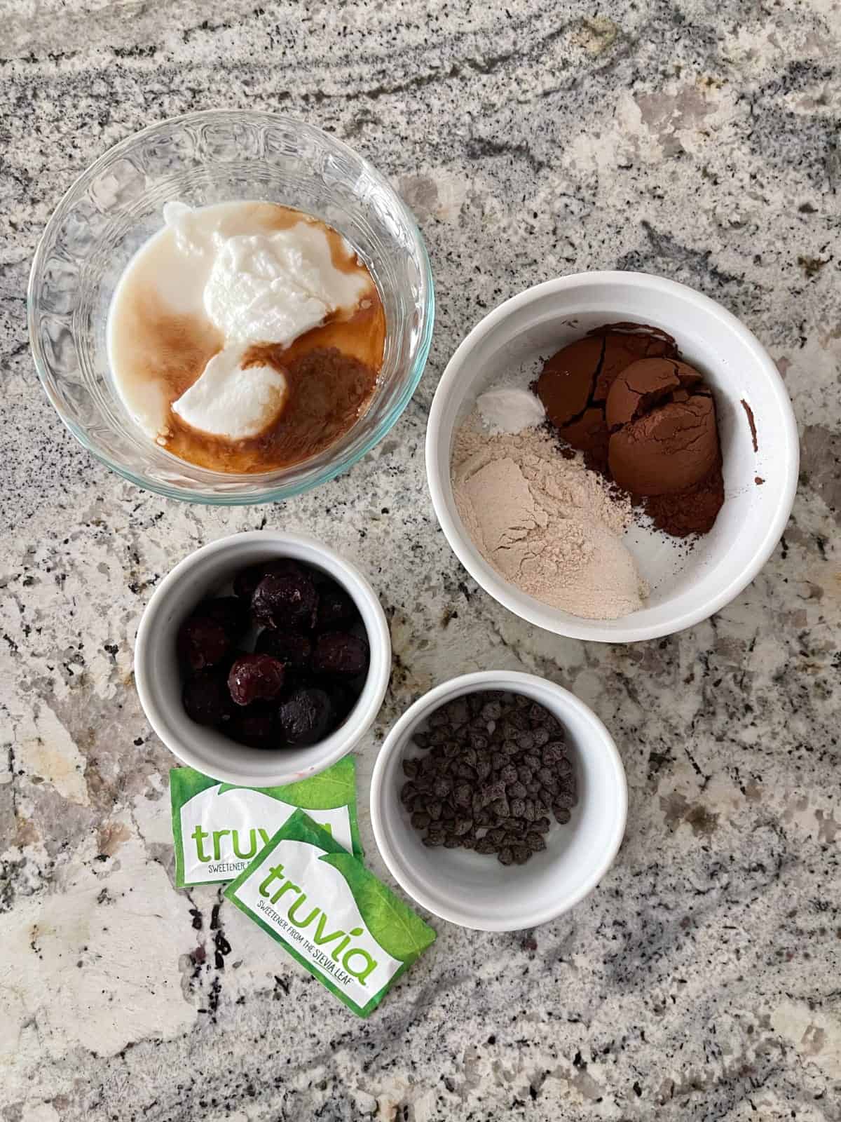 Ingredients including Truvia stevia packets, frozen cherries, chocolate chips, flour, cocoa powder, Greek yogurt, vanilla and almond milk in small bowls on granite counter.