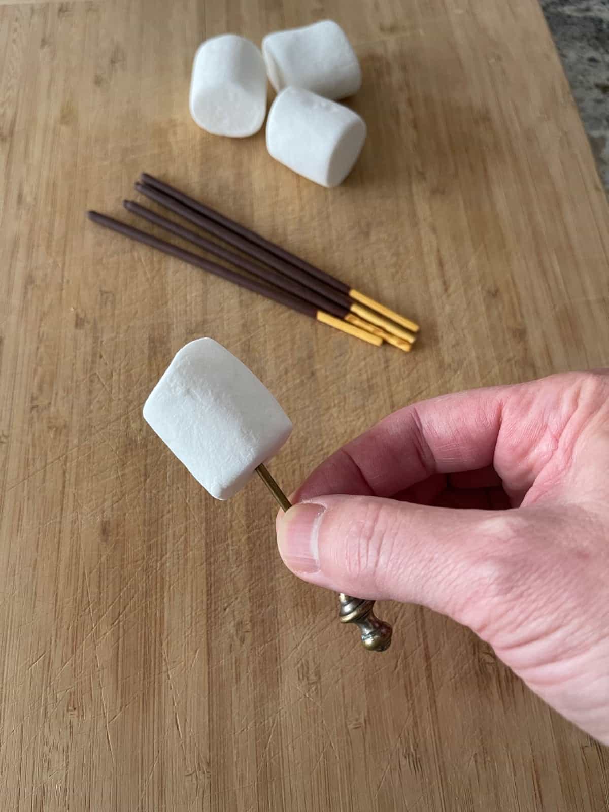 Cocktail olive skewer making hole in marshmallow with chocolate Pocky Sticks and marshmallows in background.