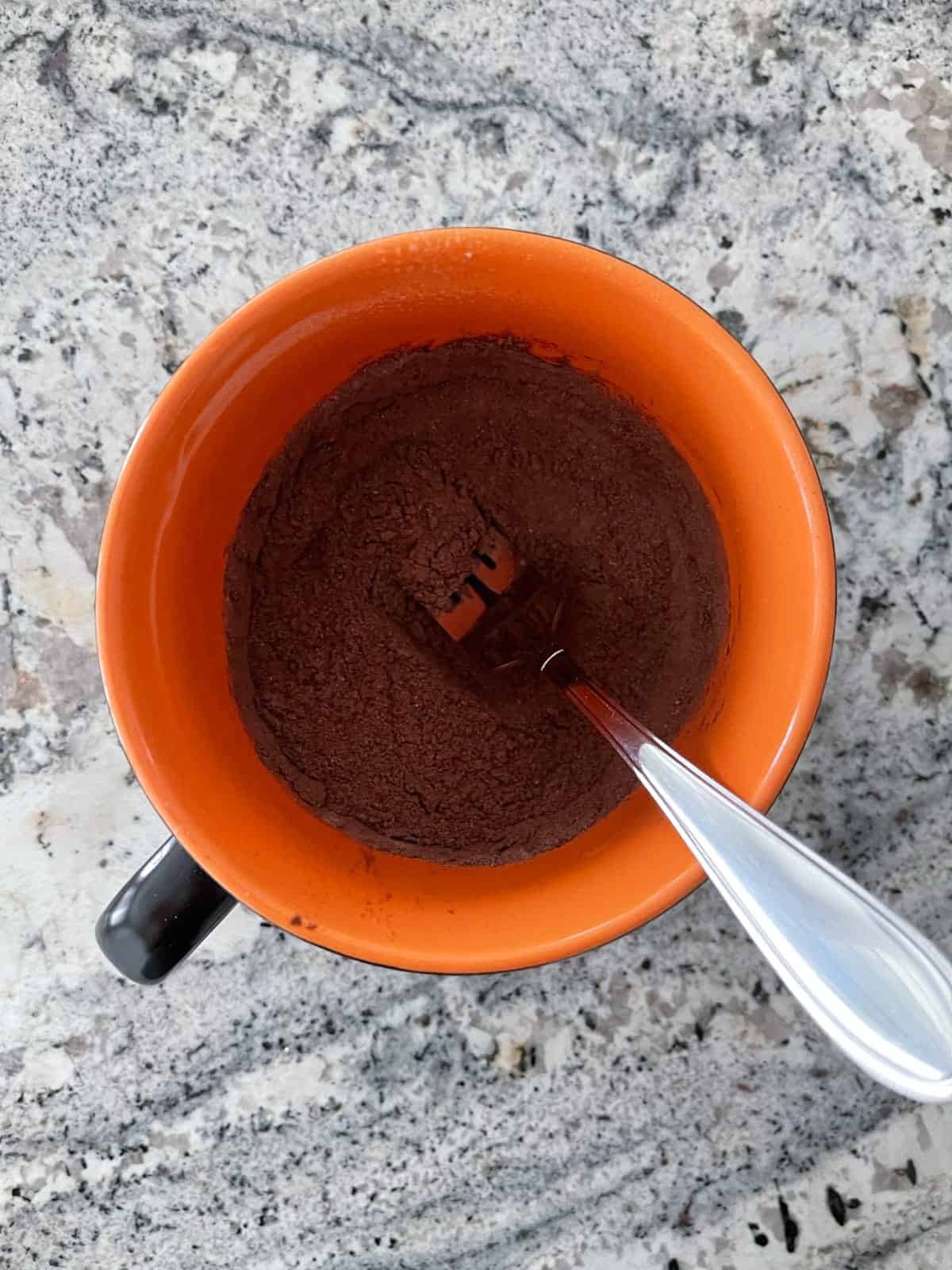 Mixing flour, cocoa powder, Truvia and baking powder with fork in orange mug.
