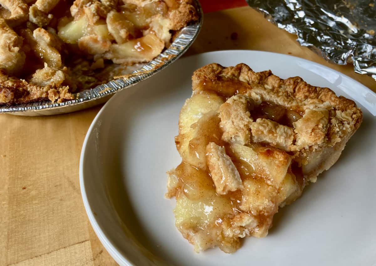 Slice of homemade apple pie on small white plate with rest of pie in background.