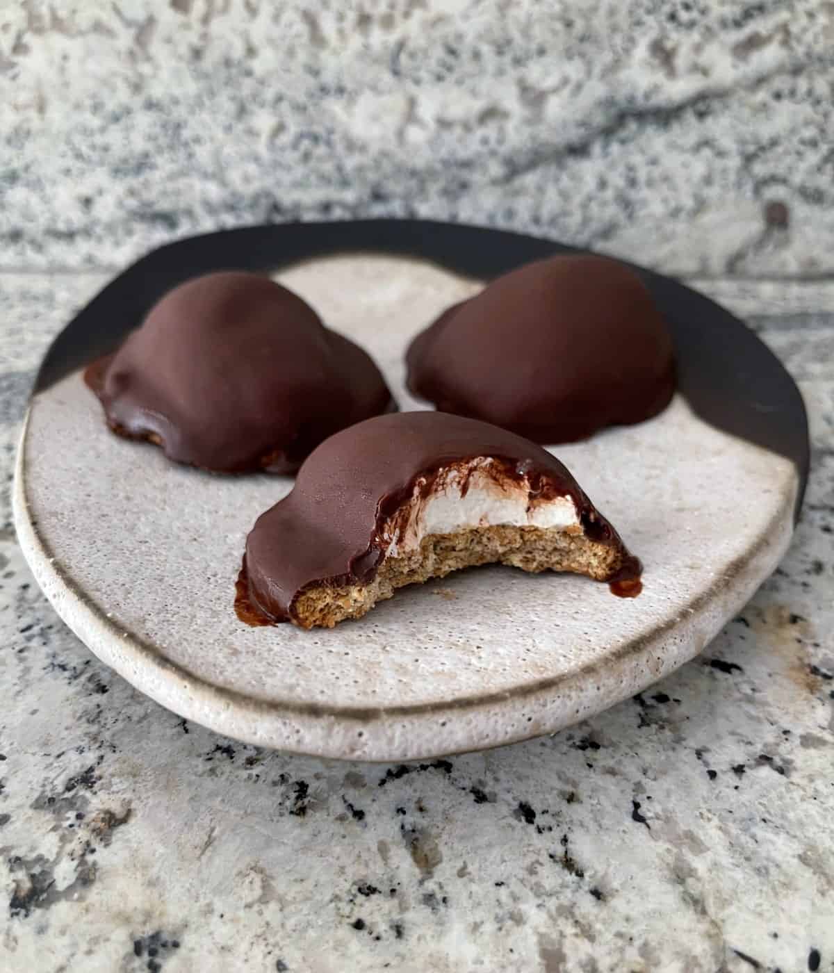 Three homemade chocolate covered marshmallow fluff cookies on ceramic plate with bite taken from one cookie.